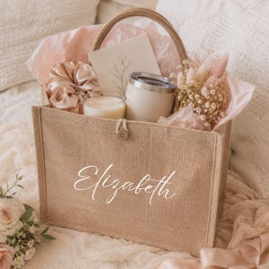 May include: A beige burlap tote bag with a handle, filled with gifts. The bag is personalized with the name "Elizabeth" in white script. Inside are a candle, tumbler, scrunchie, card, and dried flowers, all in soft, neutral tones.