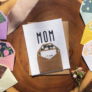 Puede incluir: Una tarjeta blanca con la palabra "MOM" en letras negras, rodeada de relieve floral, y un pequeño sobre con "I love you". Varios sobres coloridos con diseños florales están dispuestos alrededor de la tarjeta, con un pequeño ramo de flores.