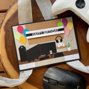 Może przedstawiać: Ręcznie robiona kartka urodzinowa z motywem gier. Karta zawiera ilustrację konfiguracji gier, balony i napis "HAPPY BIRTHDAY". Widoczny jest również biały kontroler do gier i czarna mysz komputerowa.