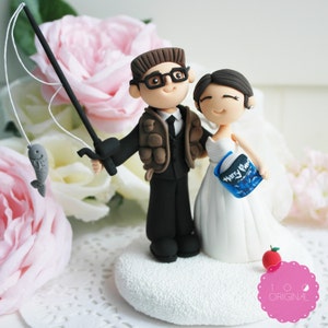 May include: A wedding cake topper featuring a bride and groom figurine. The groom is holding a fishing rod with a fish on the line. The bride is holding a book with the text "Harry Potter" on the cover. The topper is on a white heart-shaped base.