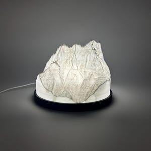 May include: A white mountain-shaped lamp with a textured surface sits on a white base and a black circular stand. The lamp is lit, casting a soft glow. The cord extends to the left.