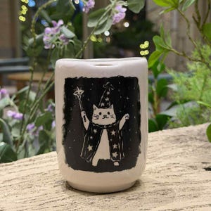 Puede incluir: Taza de cerámica blanca con una ilustración negra de un gato mago. El gato lleva un sombrero de mago, una capa y una varita, sobre un fondo de cielo estrellado. La taza tiene forma cilíndrica y un acabado liso y brillante.