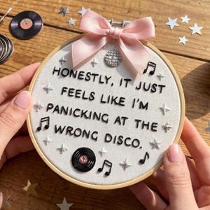 May include: Embroidered hoop art with the text "Honestly, it just feels like I'm panicking at the wrong disco." The design includes musical notes, stars, and a small disco ball charm, topped with a pink bow. A vinyl record is also embroidered.