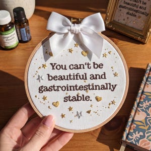 May include: An embroidered hoop art piece with the text "You can't be beautiful and gastrointestinally stable." The design features gold stars and hearts, a white bow, and a wooden frame. The piece is on a wooden surface.