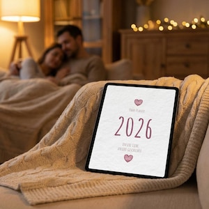 May include: A digital tablet displaying the year 2026, with the text "Paar-Planer" and "Unsere Liebe Unsere Geschichte" in a soft, mauve color. The tablet rests on a beige knitted blanket, with a couple blurred in the background.