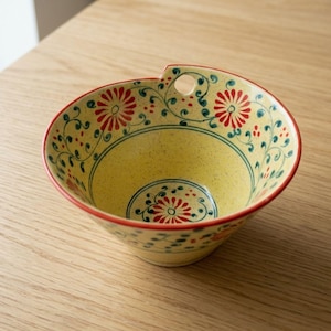 May include: A yellow ceramic bowl with a red rim and floral design. The bowl has red and green floral patterns on a yellow background. It features a small handle with a hole. The bowl is on a wooden surface.