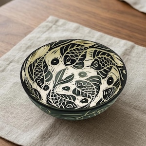 May include: A black and cream ceramic bowl with a fish design. The bowl has a detailed pattern of fish swimming in a circular arrangement. The bowl is set on a neutral linen napkin.