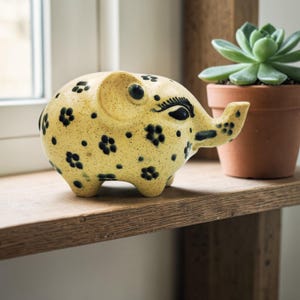 May include: A ceramic elephant figurine with a yellow base color and black floral patterns. The elephant has black eyes and a trunk. It is sitting on a wooden shelf next to a potted succulent.