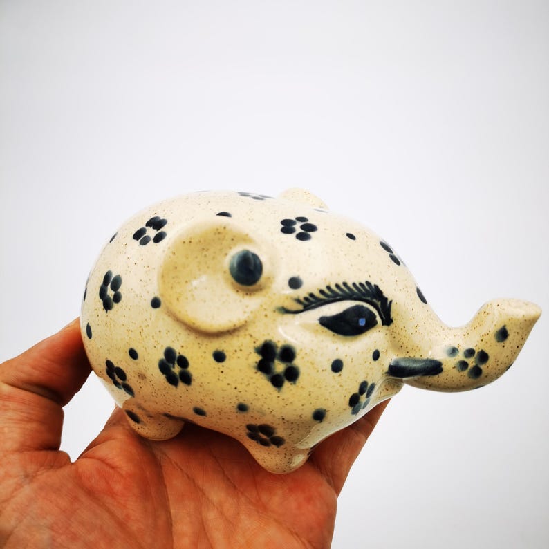 May include: A ceramic elephant figurine with a speckled beige body and dark blue floral patterns. The elephant has a detailed eye and trunk, and is held in a hand against a white background. The figurine is a decorative item.
