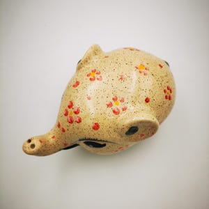 May include: A ceramic piggy bank in a light beige color, speckled with tiny brown dots. The piggy bank is decorated with red and yellow floral patterns. The pig has a long snout and black eyes.