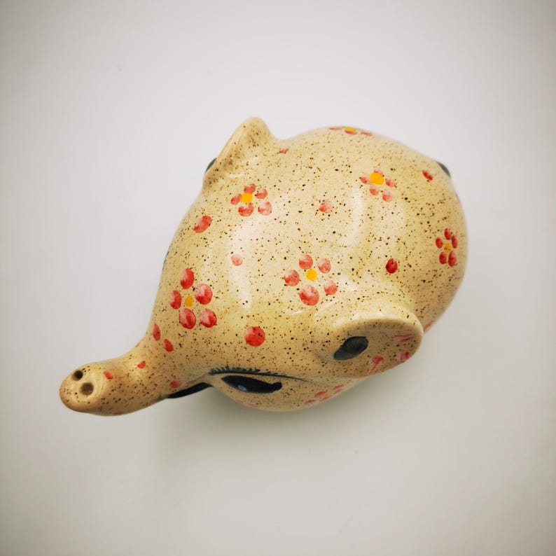 May include: A ceramic piggy bank in a light beige color, speckled with tiny brown dots. The piggy bank is decorated with red and yellow floral patterns. The pig has a long snout and black eyes.