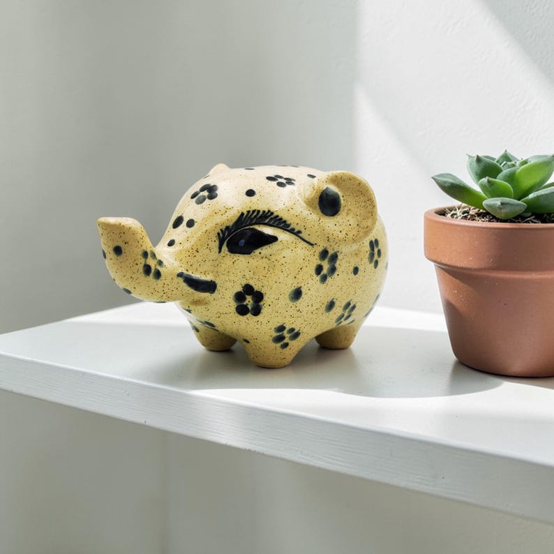 May include: A ceramic elephant planter in a light yellow color with black floral and dot patterns. The elephant has a raised trunk and small legs. A small succulent plant in a terracotta pot is next to the elephant.