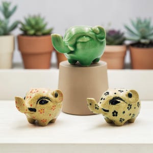 May include: Three ceramic elephant planters in the image. One is green, perched on a beige stand. The other two are tan with black and red floral patterns. Succulents in terracotta pots are in the background.