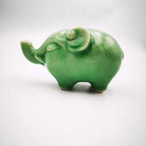 May include: A small, green ceramic elephant figurine with a glossy finish. The elephant has a detailed trunk, ears, and legs, and is standing on a white surface. The figurine is a decorative item.