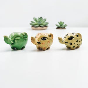 May include: Three ceramic elephant planters in green and tan with black details. The elephants have raised trunks and small ears. Two small succulent plants are in the background. The planters are suitable for small plants.