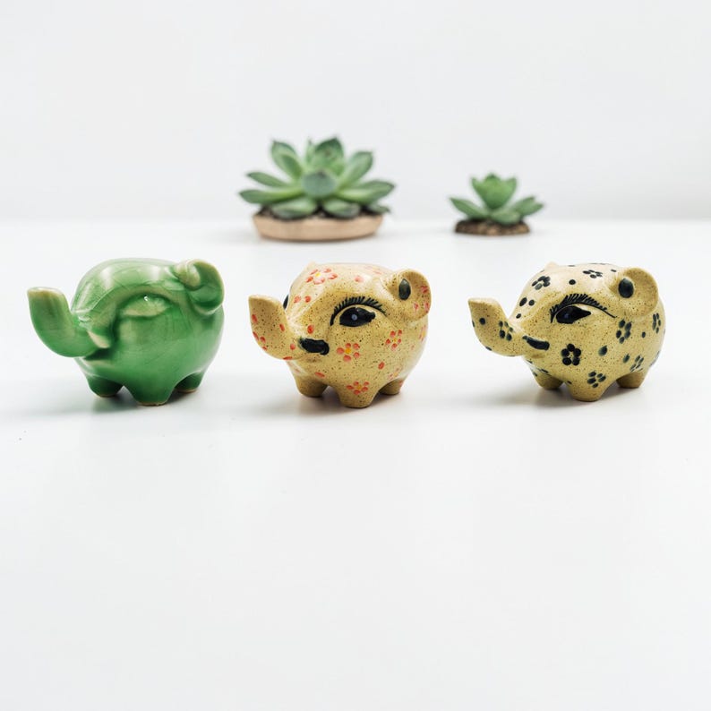 May include: Three ceramic elephant planters in green and tan with black details. The elephants have raised trunks and small ears. Two small succulent plants are in the background. The planters are suitable for small plants.