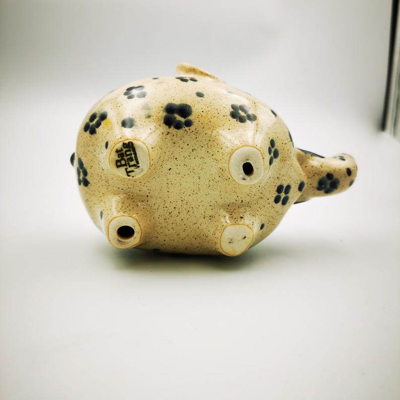 May include: A ceramic piggy bank with a speckled beige finish and dark blue floral patterns. The underside features three circular openings and the text "Bat Trang" on a small tag. The piggy bank has a small tail and ears.