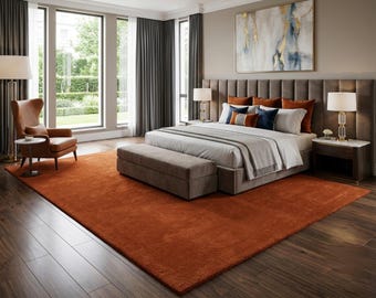 May include: A bedroom featuring a large, solid burnt orange area rug covering dark wood flooring. The room includes a bed with a grey upholstered headboard, a matching bench, and accent pillows in orange and navy blue. A leather wingback chair sits nearby.