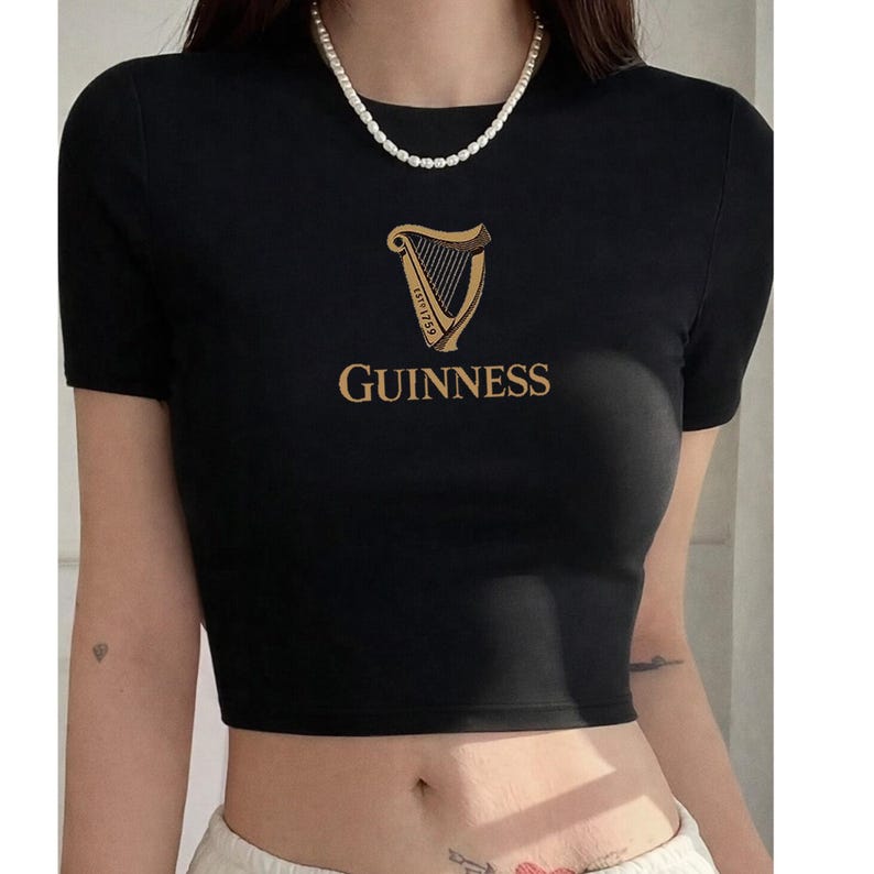May include: Black crop top with short sleeves. The gold Guinness logo, featuring a harp and the word "Guinness", is printed on the front. The top has a crew neck and is made of a soft material.