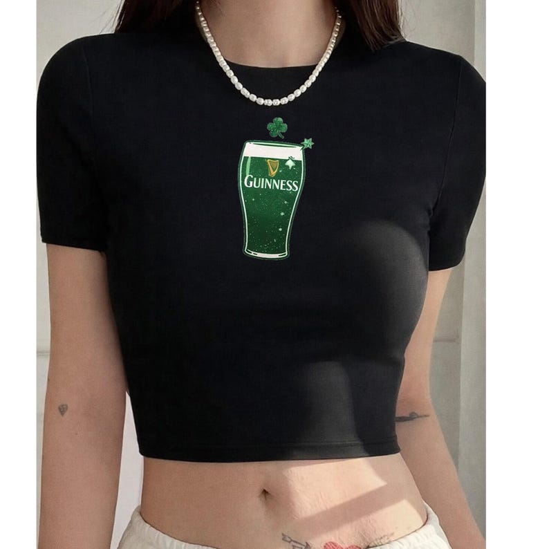 May include: Black crop top with short sleeves. The top has a green graphic of a Guinness pint glass with the Guinness logo. A small green shamrock and star are above the glass. The top is made of a soft material.