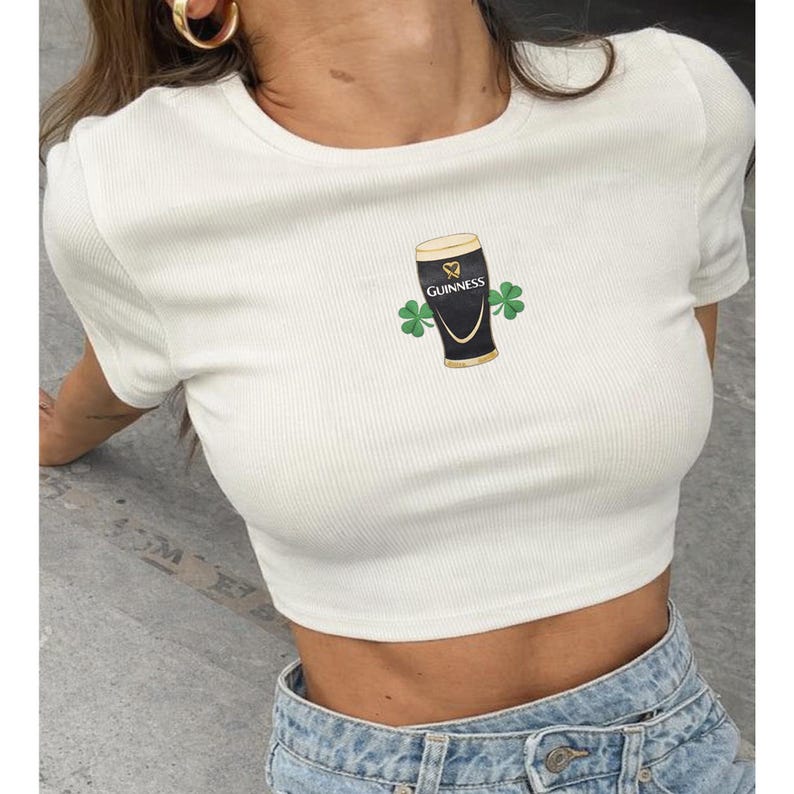 Op de afbeelding: Witte geribbelde crop top met een Guinness pint afbeelding en twee groene klavers. Het shirt heeft korte mouwen en een ronde hals. Het Guinness logo is zichtbaar op het pintglas.