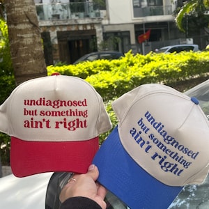 May include: Two baseball caps with the text "undiagnosed but something ain't right." One cap has a red brim and the other has a blue brim. Both caps have a beige crown.