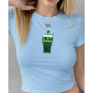 May include: Pale blue crop top with a green Guinness pint design, a shamrock, and the Guinness logo. The shirt is made of a soft material and has short sleeves. A silver necklace is worn.