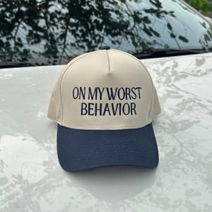 Può includere: Cappellino da baseball beige con visiera blu navy. Il cappellino presenta la scritta "ON MY WORST BEHAVIOR" in blu navy. Il cappellino è appoggiato su una superficie bianca.