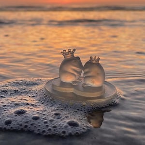 Peut inclure: Deux petites figurines de grenouilles en verre transparent portant des couronnes, assises sur une base transparente dans l'océan. L'arrière-plan montre un coucher de soleil sur l'eau, avec des teintes orange et jaune. Les grenouilles sont dans l'eau.