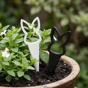 Puede incluir: Dos marcadores de plantas con forma de conejito, uno blanco y otro negro, se insertan en la tierra de una planta en maceta. Los marcadores tienen un diseño sencillo, con una silueta de cabeza de conejo en la parte superior y una base puntiaguda.