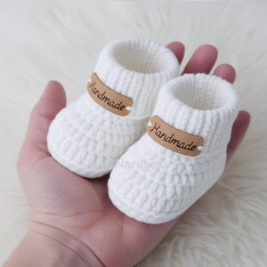 May include: A pair of handmade white crochet baby booties. Each bootie has a brown leather tag that reads "Handmade". The booties are held in a hand, and rest on a white, fluffy surface.
