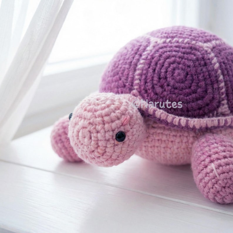 May include: A handmade crochet turtle in shades of pink and purple. The turtle has a rounded shell with a spiral design and a light pink head, feet, and tail. The eyes are black, and the turtle is sitting on a white surface.
