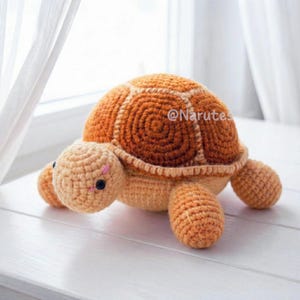 May include: A handmade crochet turtle in shades of orange and tan. The turtle has a textured shell, a round head, and four stubby legs. The eyes are small black beads, and there is a pink detail on the head. The text "@Narutes" is visible.