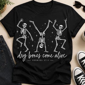 Könnte beinhalten: Ein schwarzes T-Shirt mit einer weißen Grafik von drei tanzenden Skeletten, die von kleinen Sternen umgeben sind. Unter den Skeletten steht der Text "dry bones come alive" in Schreibschrift, gefolgt von der Referenz "EZEKIEL 37:5" in Druckschrift.