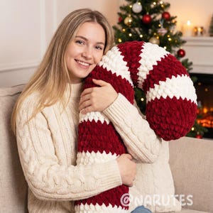 May include: A large, plush candy cane pillow in red and white stripes. The pillow is made of a soft, textured material and is curved in the shape of a candy cane. The pillow is being held by a person wearing a cream-colored sweater.