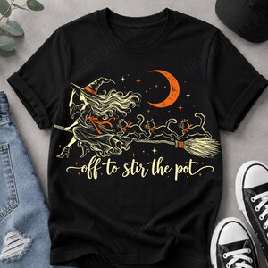 May include: A black t-shirt featuring a graphic of a witch with long hair wearing a hat and riding a broomstick with three black cats. An orange crescent moon and stars are above, with the text off to stir the pot in cursive script.