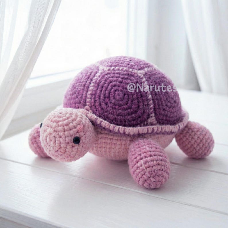 May include: A handmade crochet turtle in shades of pink and purple. The turtle has a rounded shell with a darker purple center and lighter purple edges. The head, legs, and underside are a soft pink. Black safety eyes are visible. The text "@Narutes" is visible on the shell.