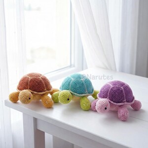 May include: Three handmade crochet turtles in a row. The turtles have different colored shells: brown, teal, and purple. The turtles have yellow, green, and pink legs and heads. The turtles are sitting on a white table.