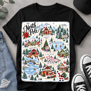 May include: A black t-shirt featuring a colourful graphic map of the North Pole. The illustration includes Santa's workshop, reindeer stables, a Christmas tree forest, and candy cane lane, with the text The North Pole printed in a black script font.