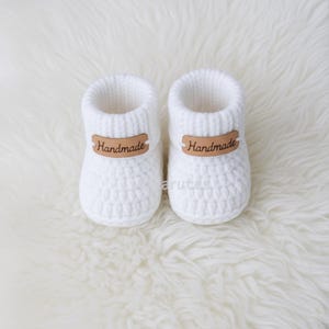 May include: A pair of white, handmade baby booties. The booties are knit with a textured stitch and feature a brown leather tag that reads "Handmade". The booties are designed to keep a baby's feet warm and cozy.