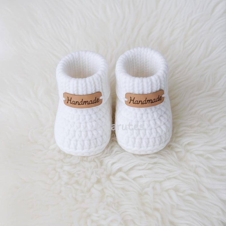 May include: A pair of white, handmade baby booties. The booties are knit with a textured stitch and feature a brown leather tag that reads "Handmade". The booties are designed to keep a baby's feet warm and cozy.