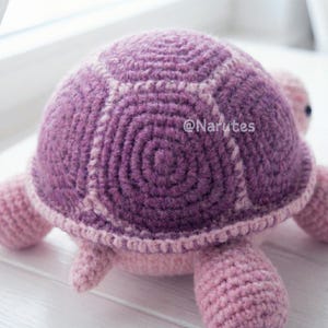 May include: A handmade crochet turtle in shades of purple and pink. The turtle's shell is a deep purple with a spiral design, and the edges are outlined in a lighter pink. The head, legs, and underside are a soft pink. The text "@Narutes" is visible on the shell.