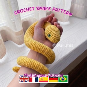 Op de afbeelding: Een gehaakte gele slang met groene ogen is om een hand gewikkeld. De afbeelding toont de tekst "CROCHET SNAKE PATTERN". De tekst "Multilingual Tutorial" met vlaggen is ook aanwezig.