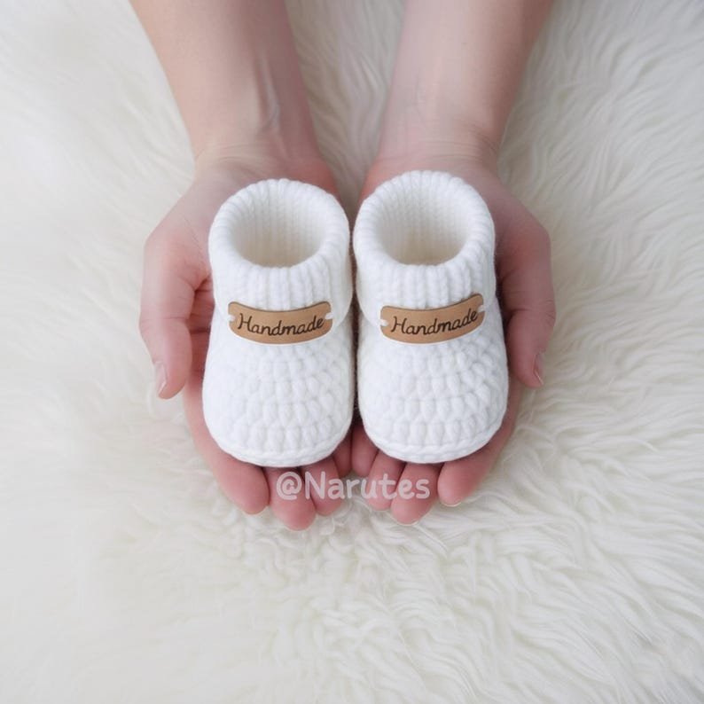 May include: A pair of white, crocheted baby booties with brown "Handmade" labels. The booties are held in open hands against a white, fluffy background. The Etsy shop name "@Narutes" is visible at the bottom.