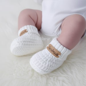 May include: A pair of white, knitted baby booties. Each bootie has a brown leather tag that reads "Handmade". The booties are on a soft, white surface.