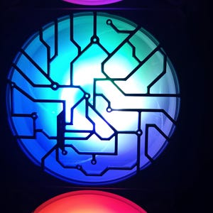 Può includere: Una ventola per computer con un design circolare con un motivo a circuito stampato. La ventola si illumina di luce blu, verde e bianca, con luce rossa e viola visibile in basso e in alto. Lo sfondo è nero.