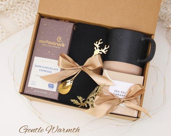 May include: A gift box filled with a black speckled mug, dark chocolate espresso bar, black socks, and sea salt caramel. The box is tied with gold ribbon and includes a gold spoon and deer ornament. The text "Gentle Warmth" is at the bottom.