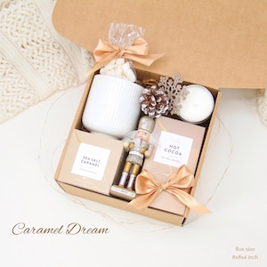 May include: A gift box labeled "Caramel Dream" filled with a white mug, marshmallows, a candle, and two boxes of cocoa. A gold nutcracker and bow add a festive touch. Box size is 8x8x4 inches.