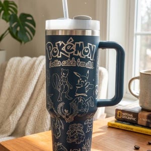 May include: A navy blue stainless steel tumbler with a white lid and straw. The tumbler features the Pokémon logo and the phrase "Gotta catch 'em all!" along with various Pokémon character illustrations. It has a large handle.