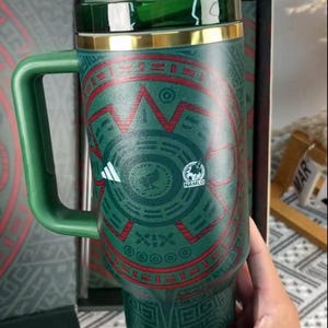Puede incluir: Vaso t&eacute;rmico verde y rojo con asa, con un dise&ntilde;o geom&eacute;trico y un borde dorado. El vaso tiene una tapa y un asa verdes, y el logotipo "NAMLO" es visible. El vaso est&aacute; en una caja negra.
